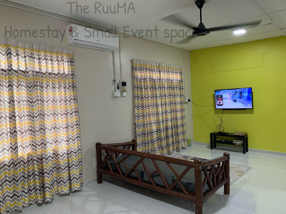 The living area features a wooden frame sofa with a cushioned seat, positioned next to a flat-screen TV mounted on the wall. Wide windows are adorned with patterned curtains, allowing natural light to fill the space. A cool, light tile floor complements the cheerful green accent wall.