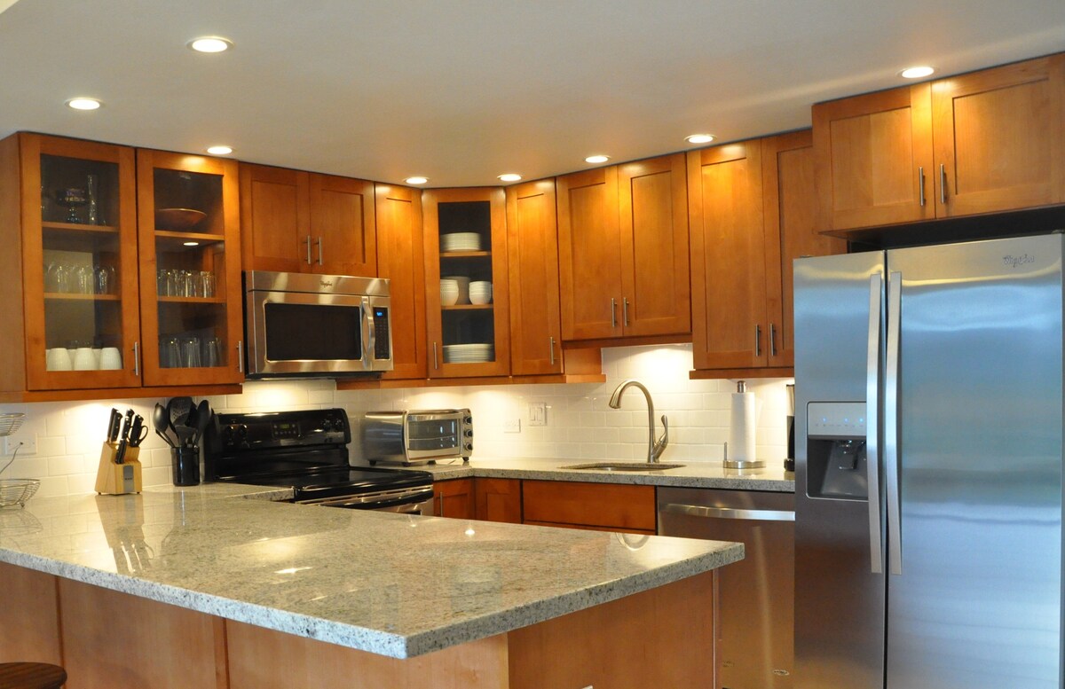 A newly remodeled kitchen is displayed, featuring rich wooden cabinets and sleek granite countertops. Stainless steel appliances, including a refrigerator, microwave, and dishwasher, complement the modern design. Glass-front cabinets showcase dishware, while under-cabinet lighting enhances the overall brightness of the space.