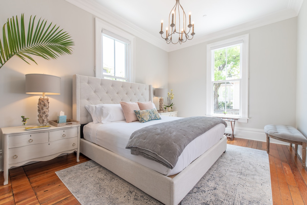 The bedroom features a spacious bed with soft linens and a decorative throw. A stylish chandelier hangs above, while large windows allow natural light to illuminate the space. A palm plant and elegant dresser add a touch of greenery and warmth to the room.