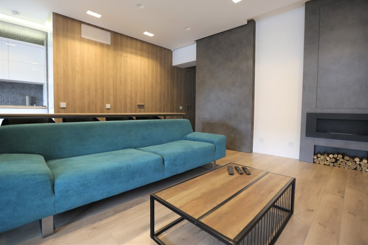 A modern living area features a large turquoise sofa facing a sleek coffee table. A bio-fireplace integrates into the wall, enhanced by a wooden accent wall. The kitchen area is visible in the background, highlighting a minimalist aesthetic with subtle lighting.