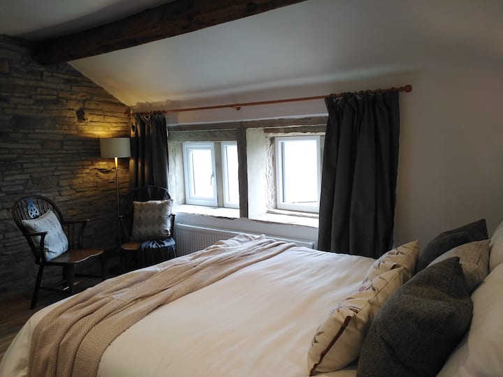 Weaverova dovolenková chata, Ripponden. Hlavná spálňa s výhľadom na priestor na posedenie pred pôvodnými kamennými stenami. Pohodlné posedenie vedľa kamenných mulionových okien vám umožní obdivovať nepretržitý výhľad na údolie Ryburn.