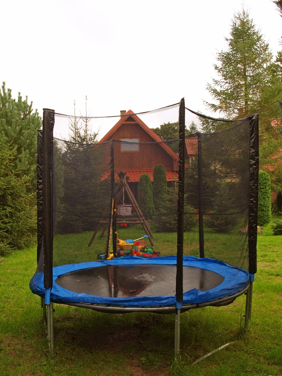 A trampoline is surrounded by a net enclosure, situated on a grassy area. In the background, a wooden house with a sloped roof is framed by tall trees. Several toys can be seen near the trampoline, hinting at a space designed for family-friendly outdoor activities.