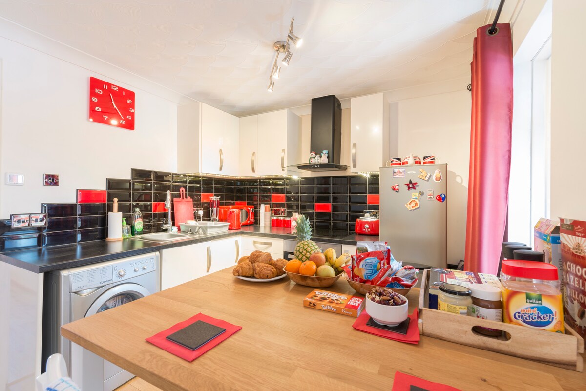 The kitchen features modern appliances and a spacious countertop. A selection of fruits, pastries, and snacks is arranged attractively, with red accents throughout the cabinets and decor. Bright natural light enters through the window, enhancing the vibrant atmosphere of the open plan space.