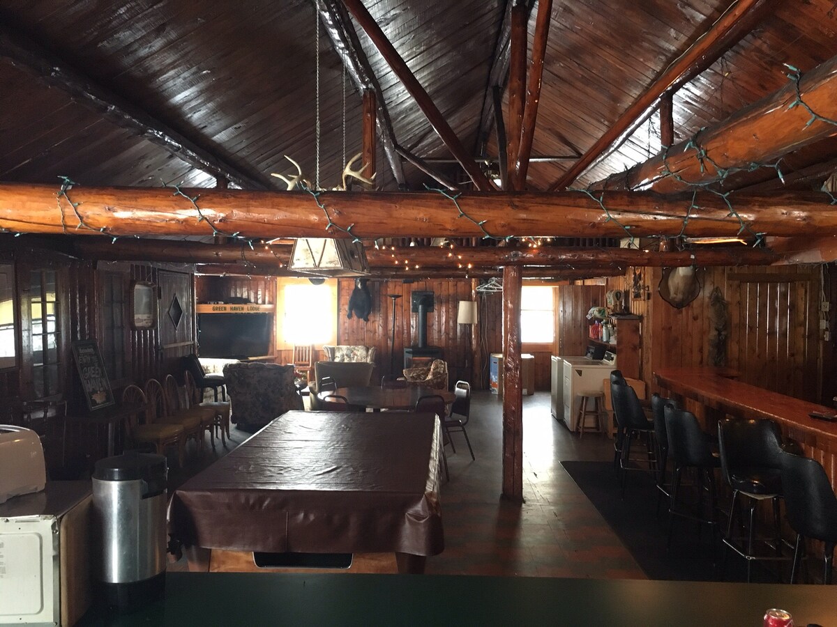 The spacious interior features original log walls and a high wood-beamed ceiling. A bar area with seating and a food prep space is visible, along with a cozy seating arrangement. Decorative lighting hangs above, and a large screen can be seen in the background.