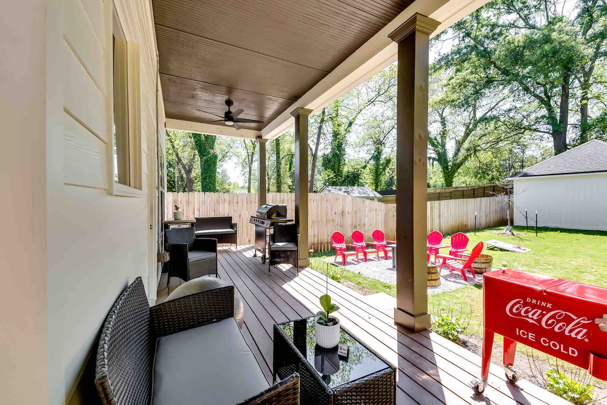 A covered patio area is furnished with black wicker seating and a glass-top table. A grill is positioned nearby, while the spacious backyard features a grassy area with lawn games and colorful lounge chairs. Fence lines the perimeter, ensuring privacy.