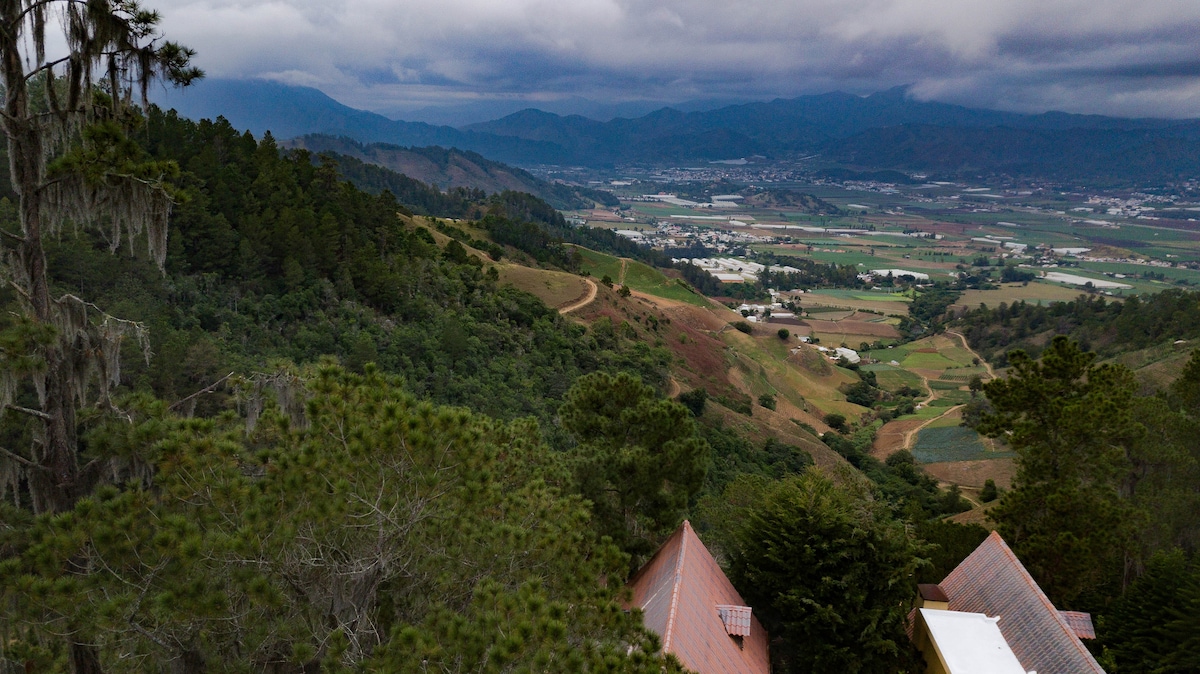 A sweeping view of the Constanza valley is captured, showcasing layered hills and lush green farmland. The rugged mountain tops are visible in the distance under a cloudy sky. Rustic rooftops peek through the surrounding trees, hinting at the serene elevation of the area.