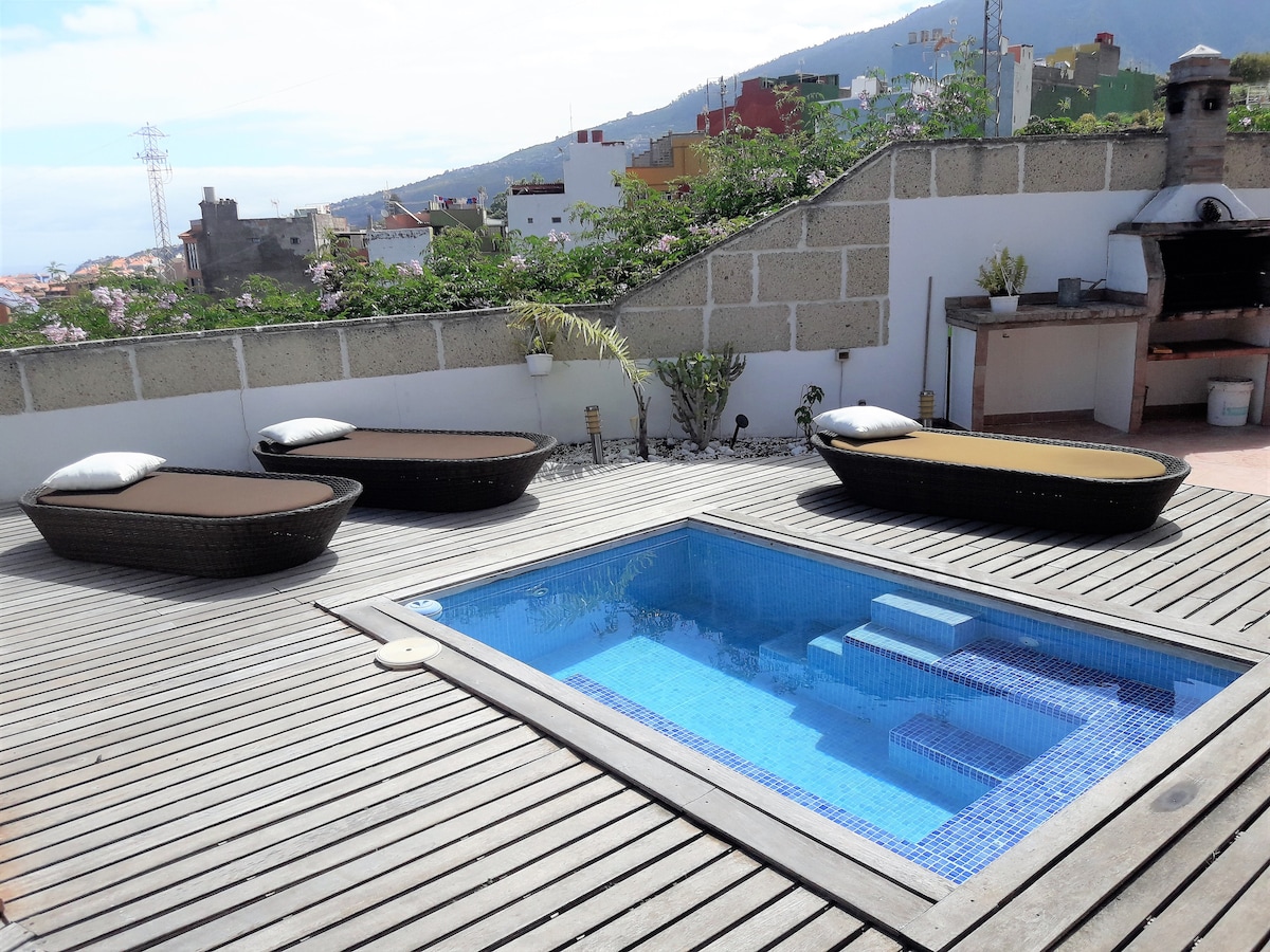 A sunlit terrace features a small private pool surrounded by a wooden deck. Three lounge chairs are positioned nearby, offering a space for relaxation. In the background, colorful buildings dot the hillside, with a view of greenery and mountains.