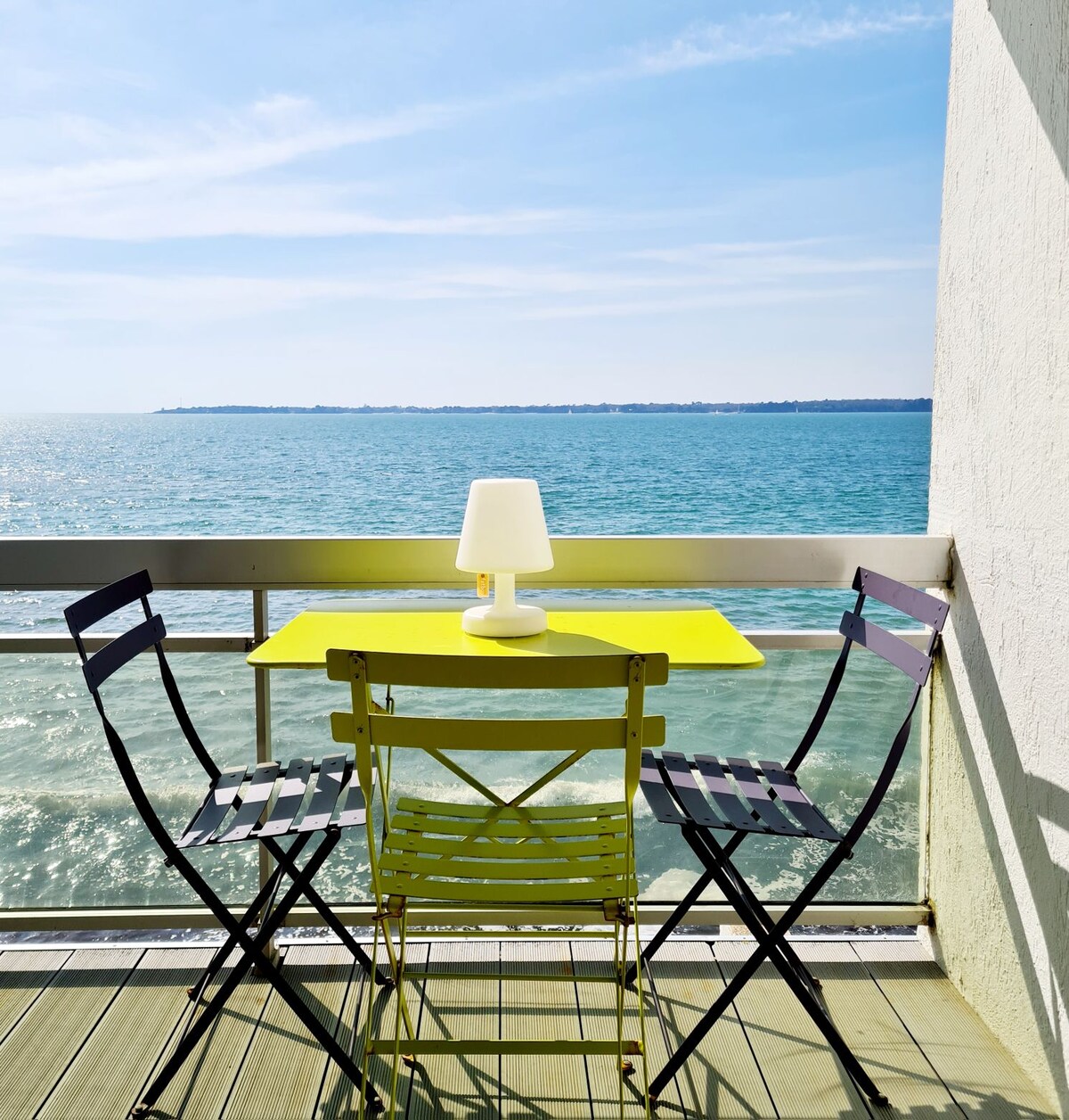 A small balcony features a bright yellow table and two folding chairs, positioned to enjoy views of the sea. A lamp sits on the table, while the blue water and distant shoreline are visible under a clear sky.