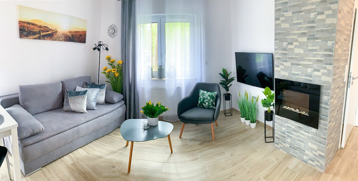 A modern living area features a comfortable gray sofa adorned with decorative pillows. A round glass coffee table sits in the center, surrounded by lush green plants. Natural light filters through the window, illuminating the stylish fireplace and accent wall.