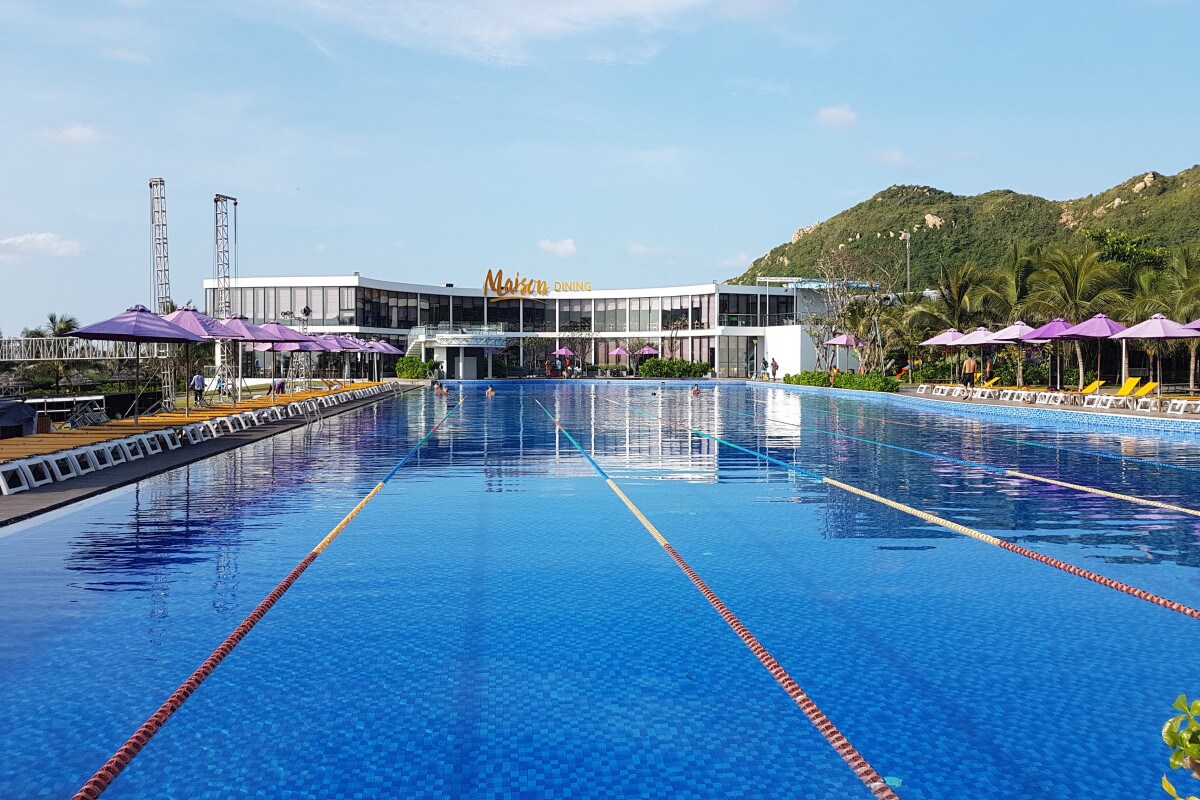 A large communal pool reflects the sky, bordered by purple umbrellas providing shade. The pool area features a spacious deck with lounge chairs arranged for relaxation. The backdrop includes a modern building and lush green hills, enhancing the resort's inviting setting.