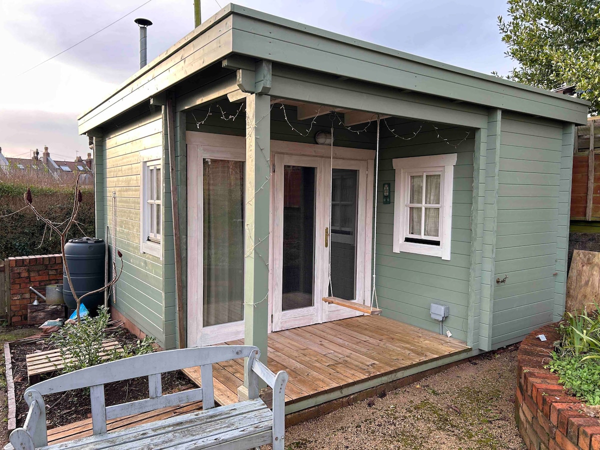 The exterior of a green garden cabin features large glass doors and windows, allowing natural light to brighten the space. A wooden deck provides access, while a simple bench and small garden area with plants can be observed nearby.