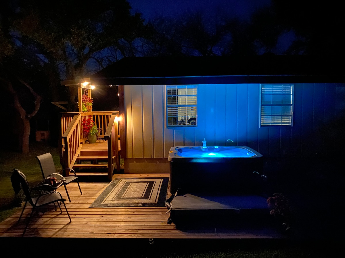 The image captures a private deck at night, featuring a hot tub illuminated in soft blue light. A chair is positioned beside the tub, and a small table sits nearby. The exterior of the suite is visible, with warm lighting accentuating the entrance and nearby foliage.