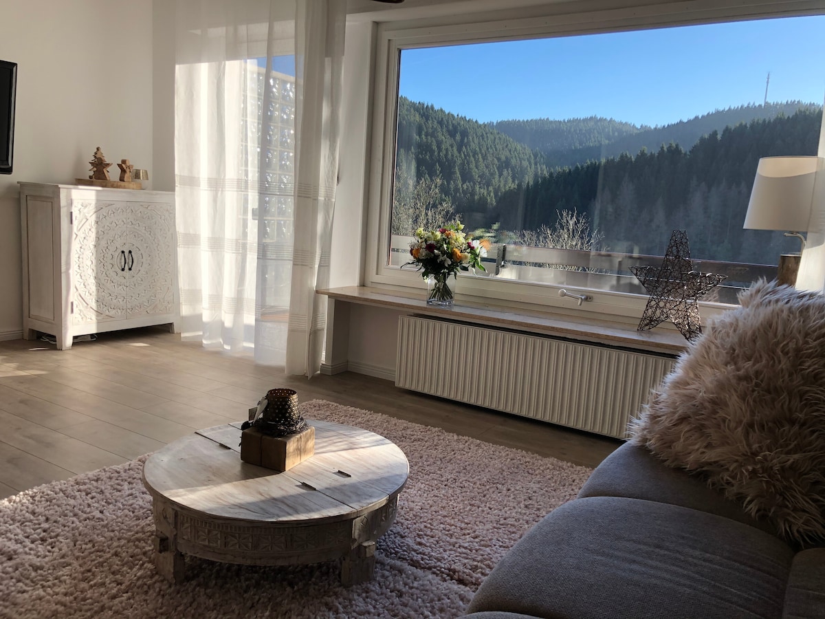 The living area features a large window revealing a panoramic view of the surrounding green mountains. A cozy seating arrangement includes a plush sofa and a decorative coffee table. Natural light floods the space, complemented by a small bouquet of flowers and a soft rug underfoot.