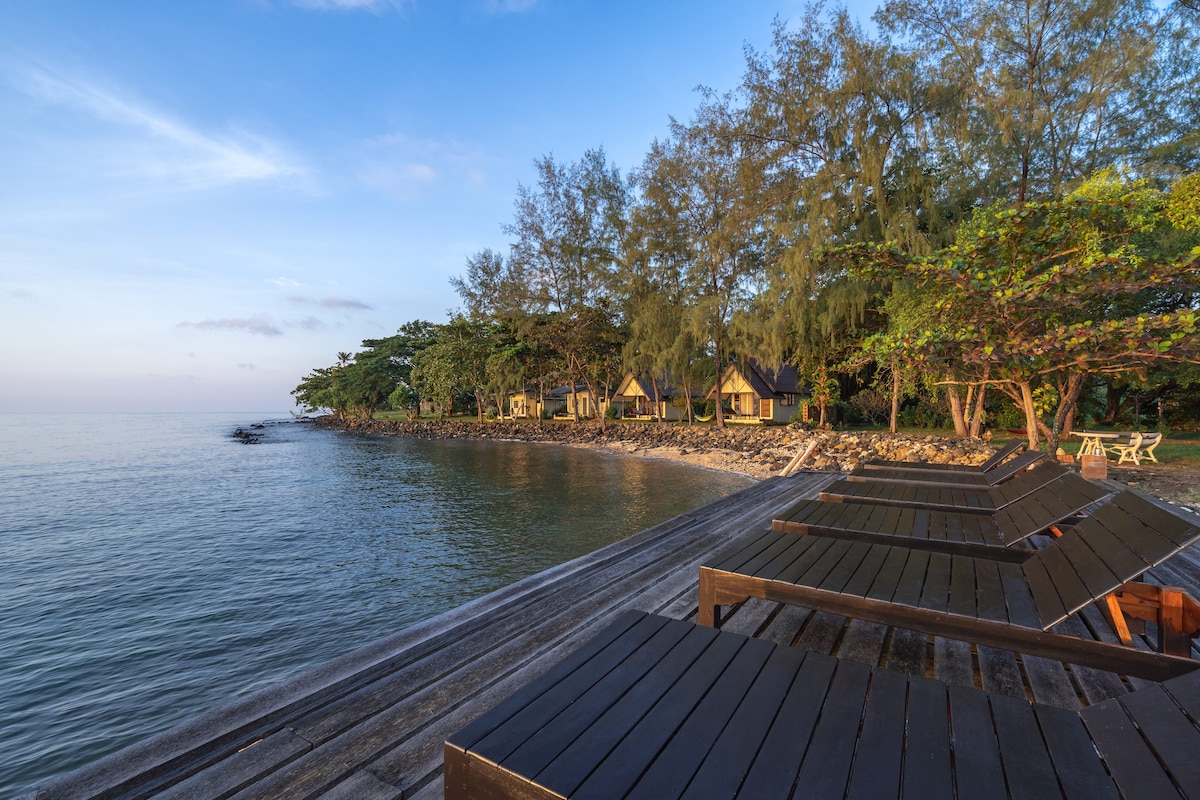 A calm seaside view features wooden loungers positioned along a boardwalk leading to the water's edge. Lush greenery and trees are visible in the background, with several colorful bungalows peeking through the foliage, creating a serene coastal atmosphere.