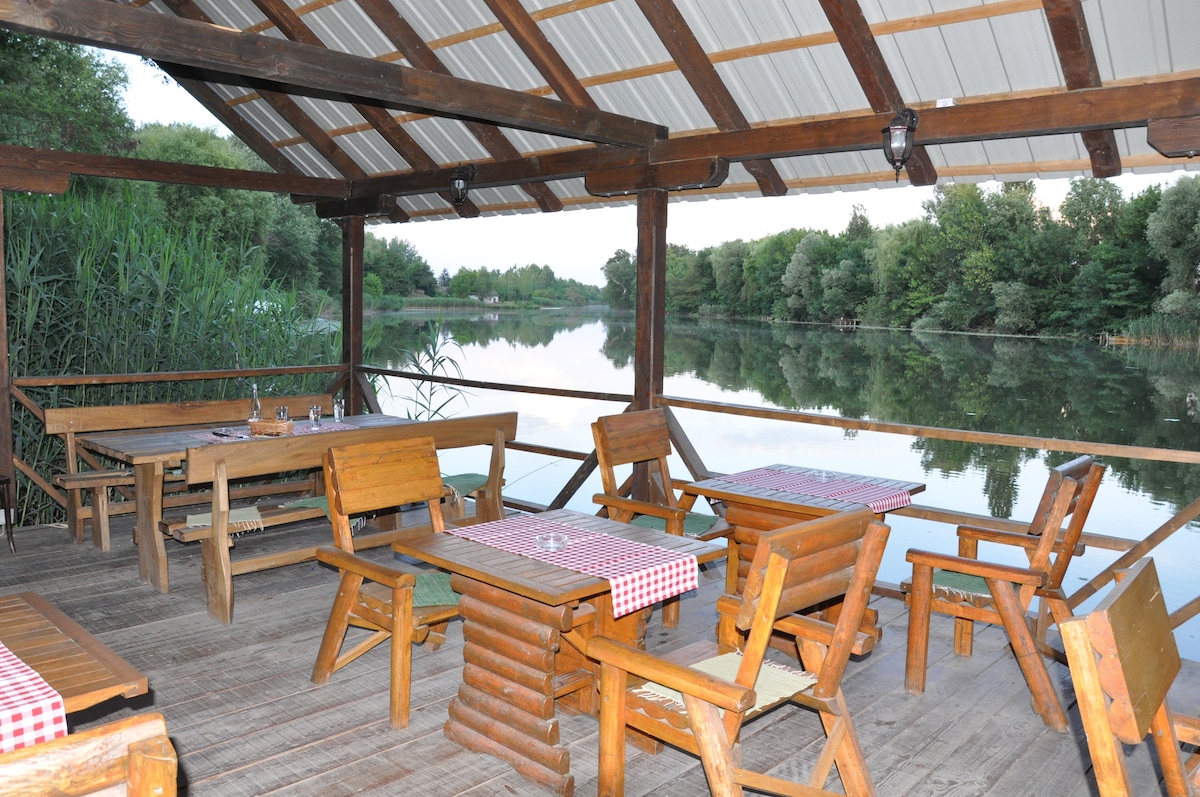 A covered terrace overlooks a tranquil body of water, surrounded by lush greenery. Wooden tables and chairs are arranged neatly, with checkered tablecloths adding a rustic touch. The serene landscape reflects in the calm water, creating a peaceful atmosphere.