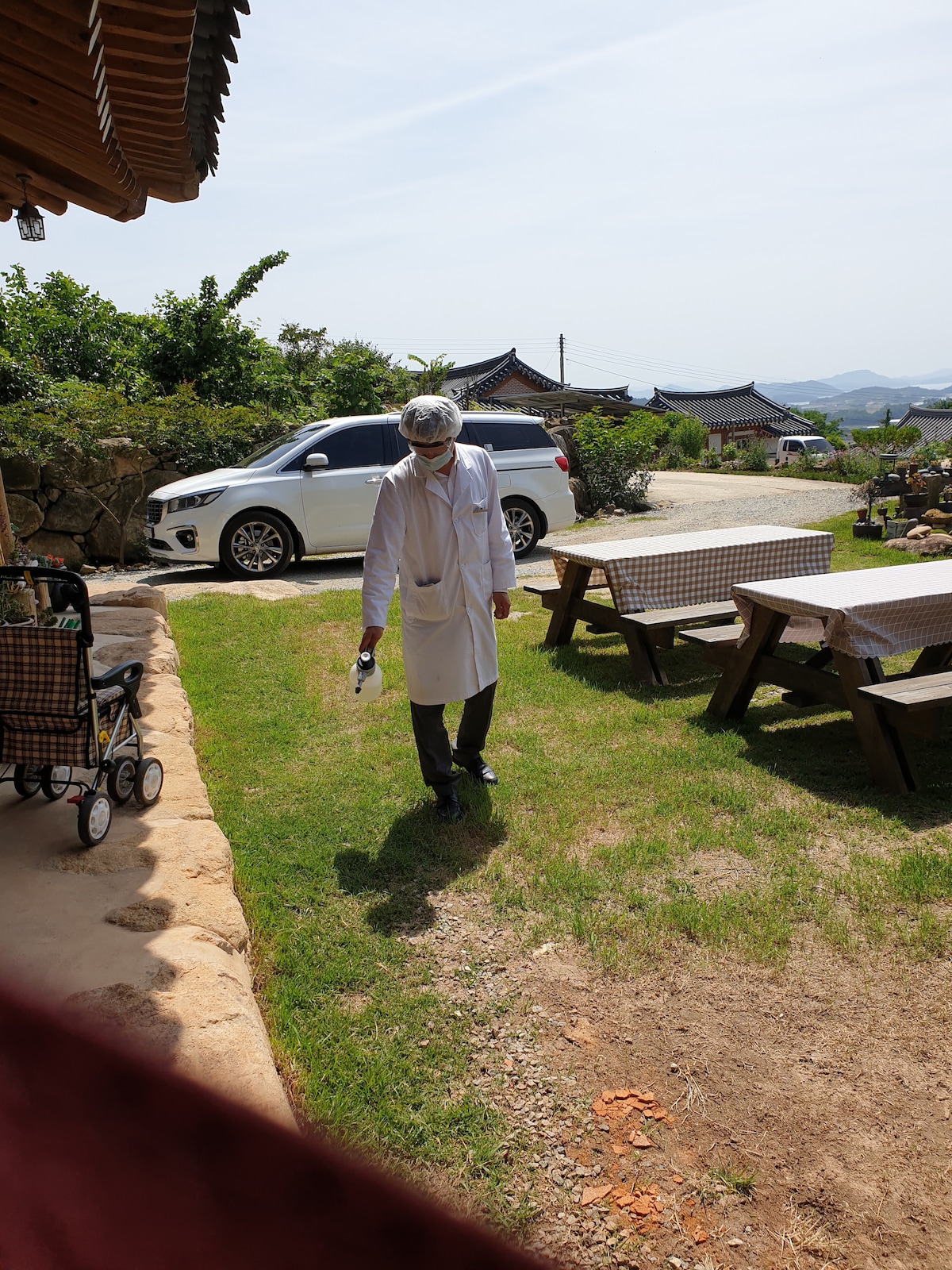 A bright outdoor space is visible, featuring green grass and wooden picnic tables. A person wearing a white apron walks across the lawn, with a vehicle parked nearby. Traditional Korean houses can be seen in the background, set against a clear sky.