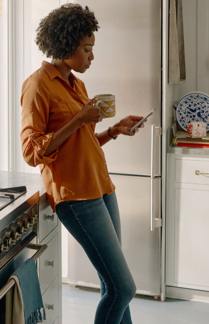 A person leans against the counter in a kitchen, holding a mug and reading something on their cell phone.