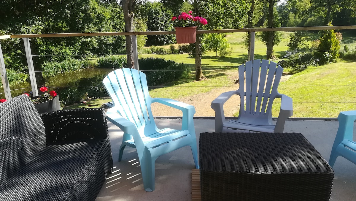 A spacious terrace features a mix of seating options, including light blue and white chairs. A dark wicker coffee table is positioned at the center. Surrounding greenery adds to the tranquil ambiance, with flower boxes hanging and vibrant blooms visible nearby.