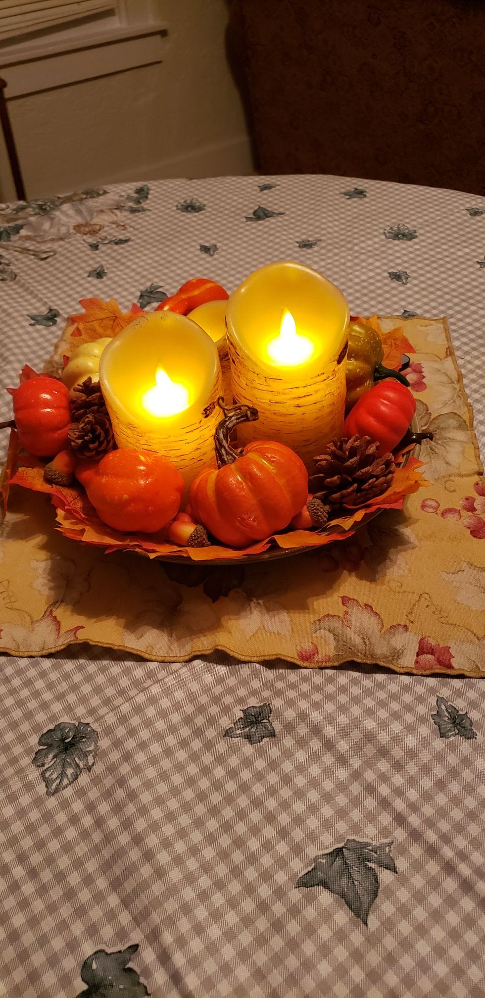 A decorative centerpiece features two glowing candles surrounded by small orange pumpkins and pine cones. The arrangement sits on a floral-patterned tablecloth, complemented by a leaf-shaped tray underneath, enhancing the seasonal ambiance.