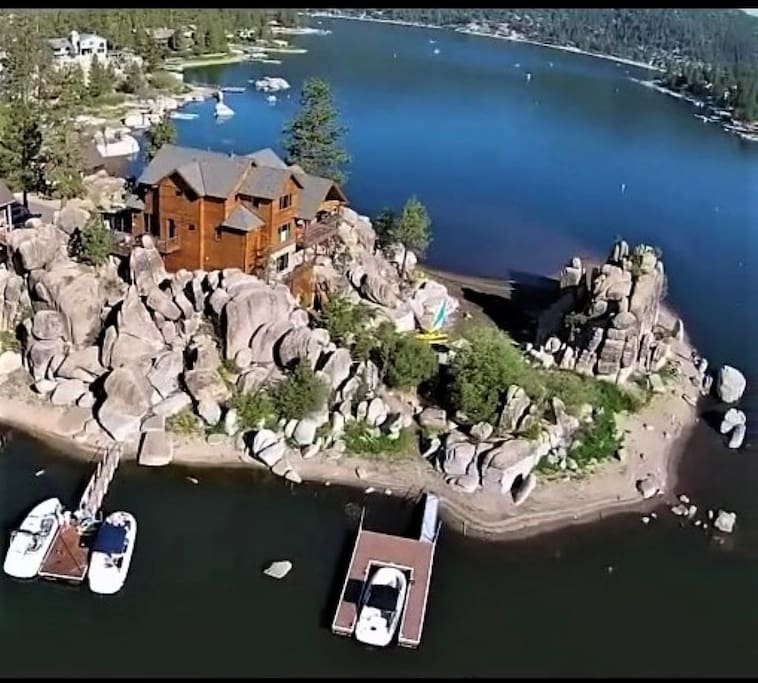 An aerial view showcases the Boulder Cove home nestled among large boulders. The property features a private dock and sandy beach with two boats moored nearby, while the scenic waters of Boulder Bay and surrounding trees are visible in the background.