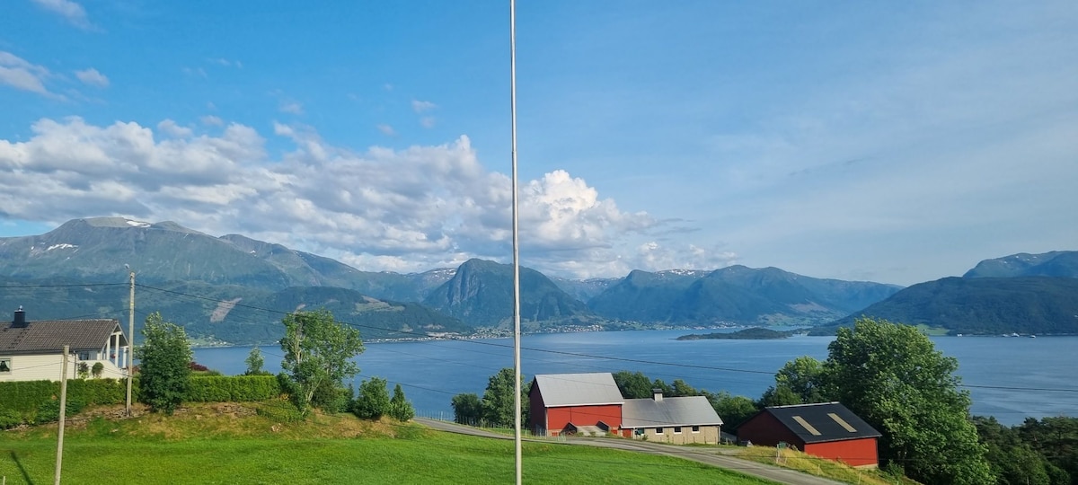 A stunning view showcases the expansive landscape, featuring the Hardangerfjorden and the surrounding mountains. The scene is complemented by lush greenery and traditional red farm buildings nestled against the hillside, set against a clear blue sky with scattered clouds.