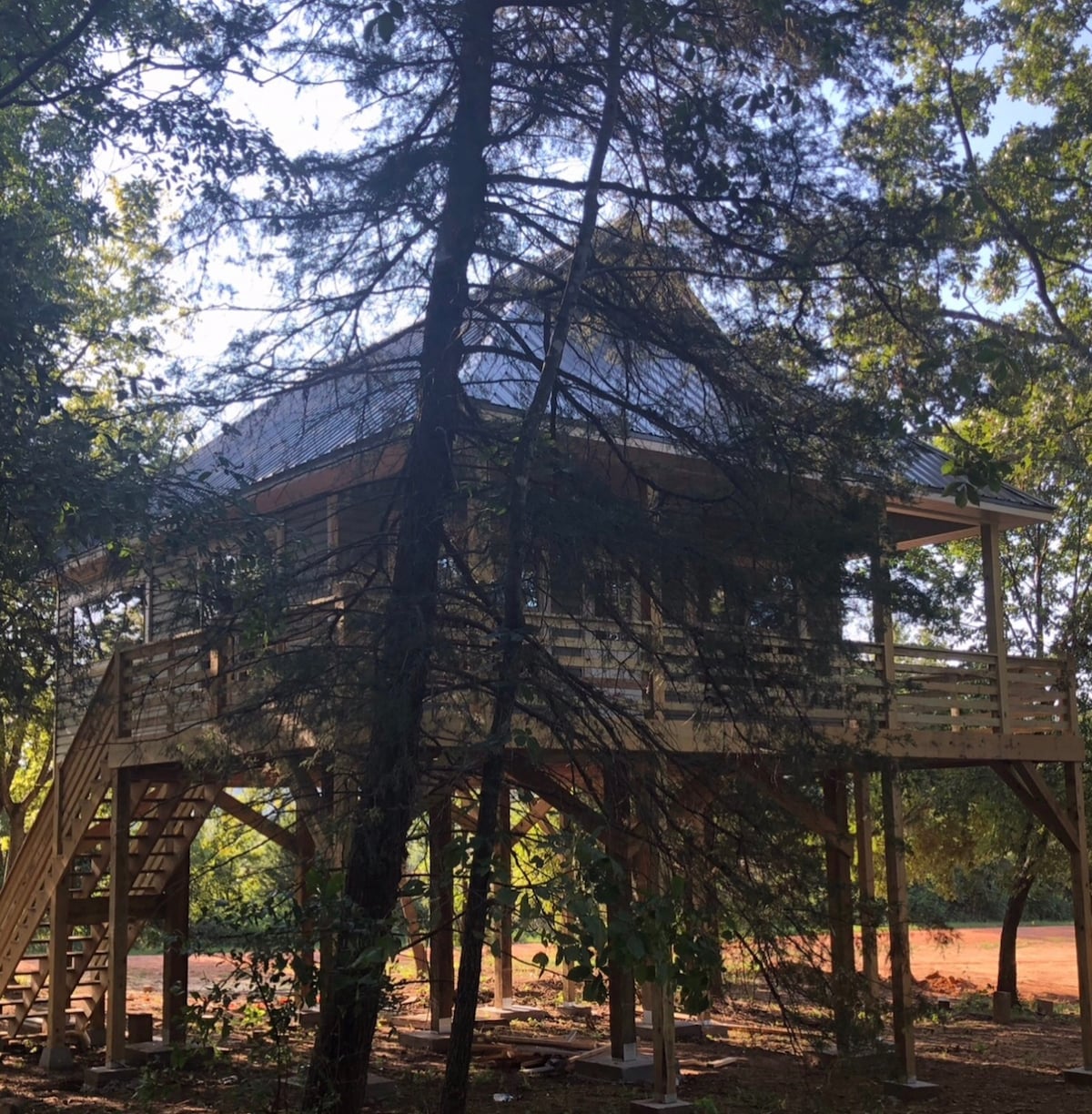 A treehouse is elevated on wooden stilts, surrounded by tall trees. A staircase leads to the main entrance, while the structure features a sloped roof and a spacious deck. The area is dappled with sunlight filtering through the foliage, creating a serene natural setting.