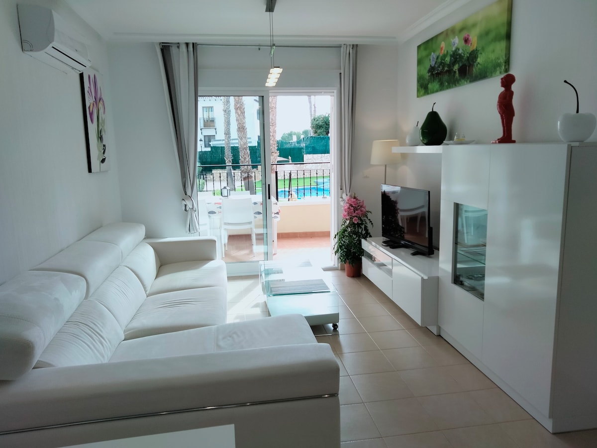 A modern living area features a light-colored sectional sofa, complemented by a glass coffee table. A flat-screen television is mounted on a sleek white console. Large glass doors provide access to a balcony, allowing natural light to fill the space.