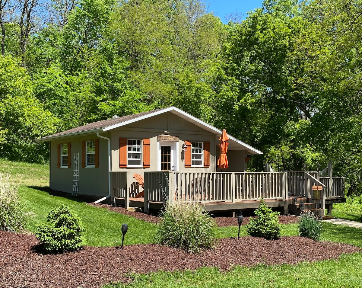 A charming cottage is set amidst lush greenery, featuring a large deck with an umbrella and wooden railings. The exterior showcases orange shutters and windows, surrounded by neatly landscaped plants and mulch. Sunlight filters through the trees, creating a serene atmosphere.