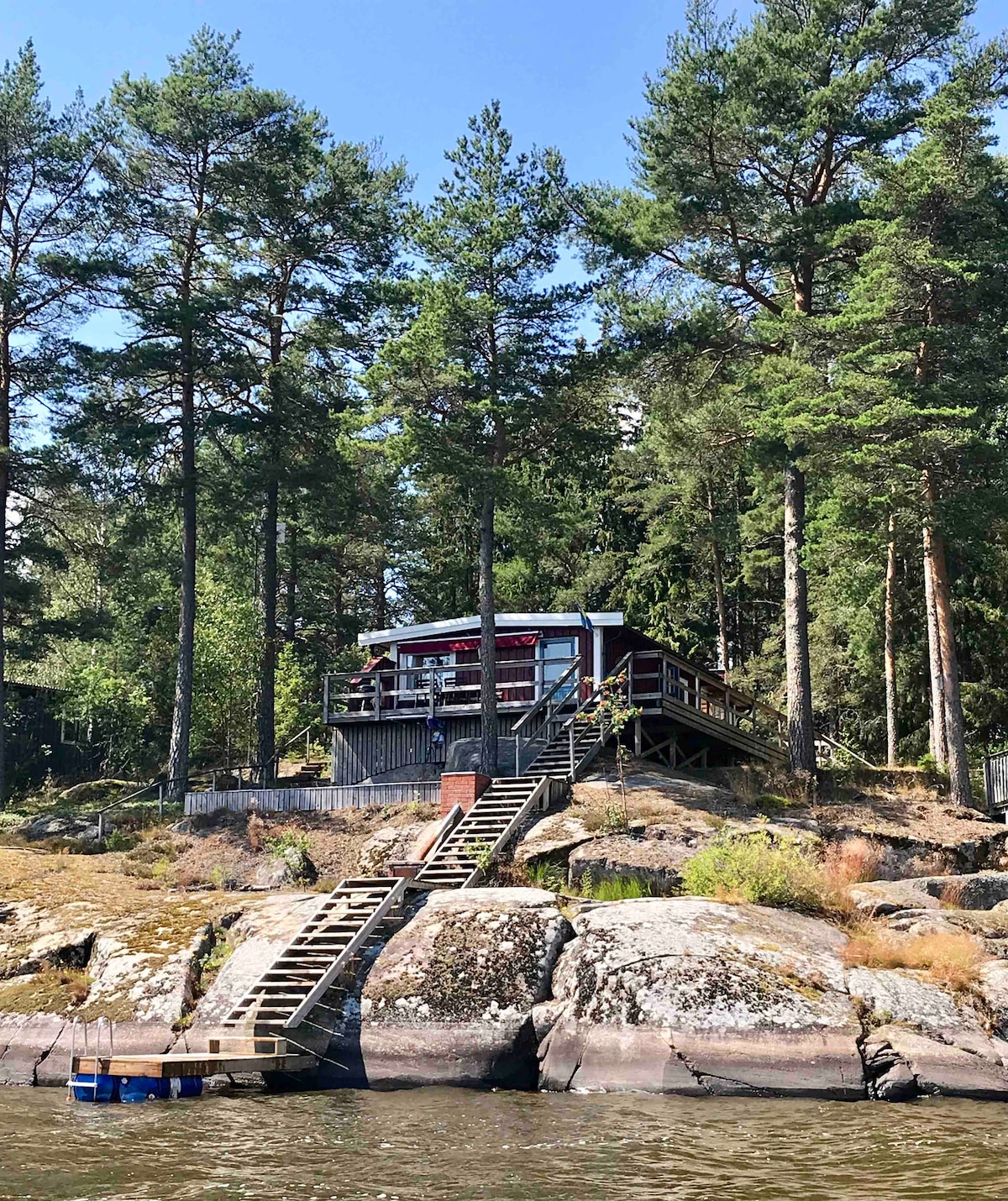A summer cottage is perched on a rocky shore, surrounded by tall trees. A wooden staircase leads down to the water from a spacious deck, creating an inviting connection to nature. The area is tranquil, with a backdrop of lush greenery.