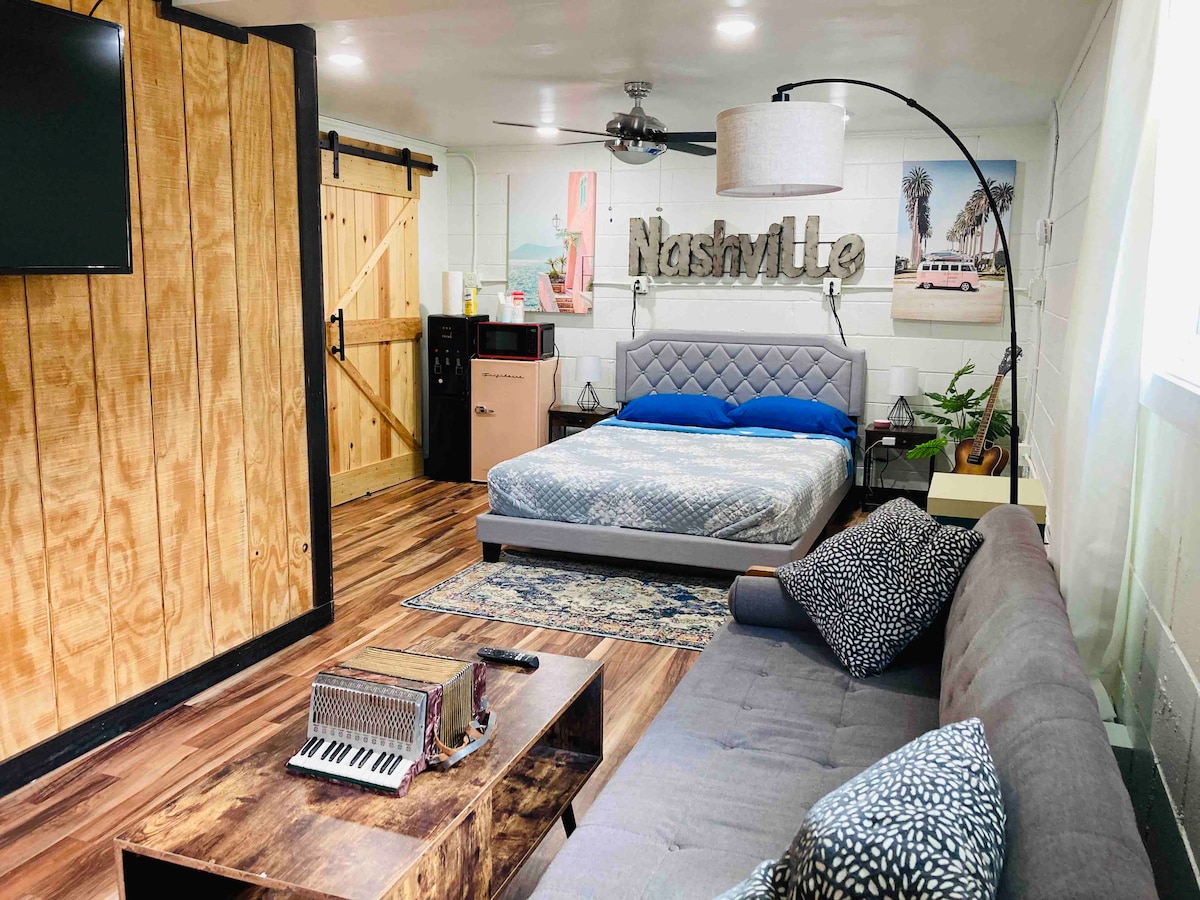 A private studio features a comfortable queen bed with blue bedding and a cozy seating area. A flat-screen TV is mounted on the wall, and a decorative 'Nashville' sign adds character. A compact fridge and a potted plant are visible in the background.