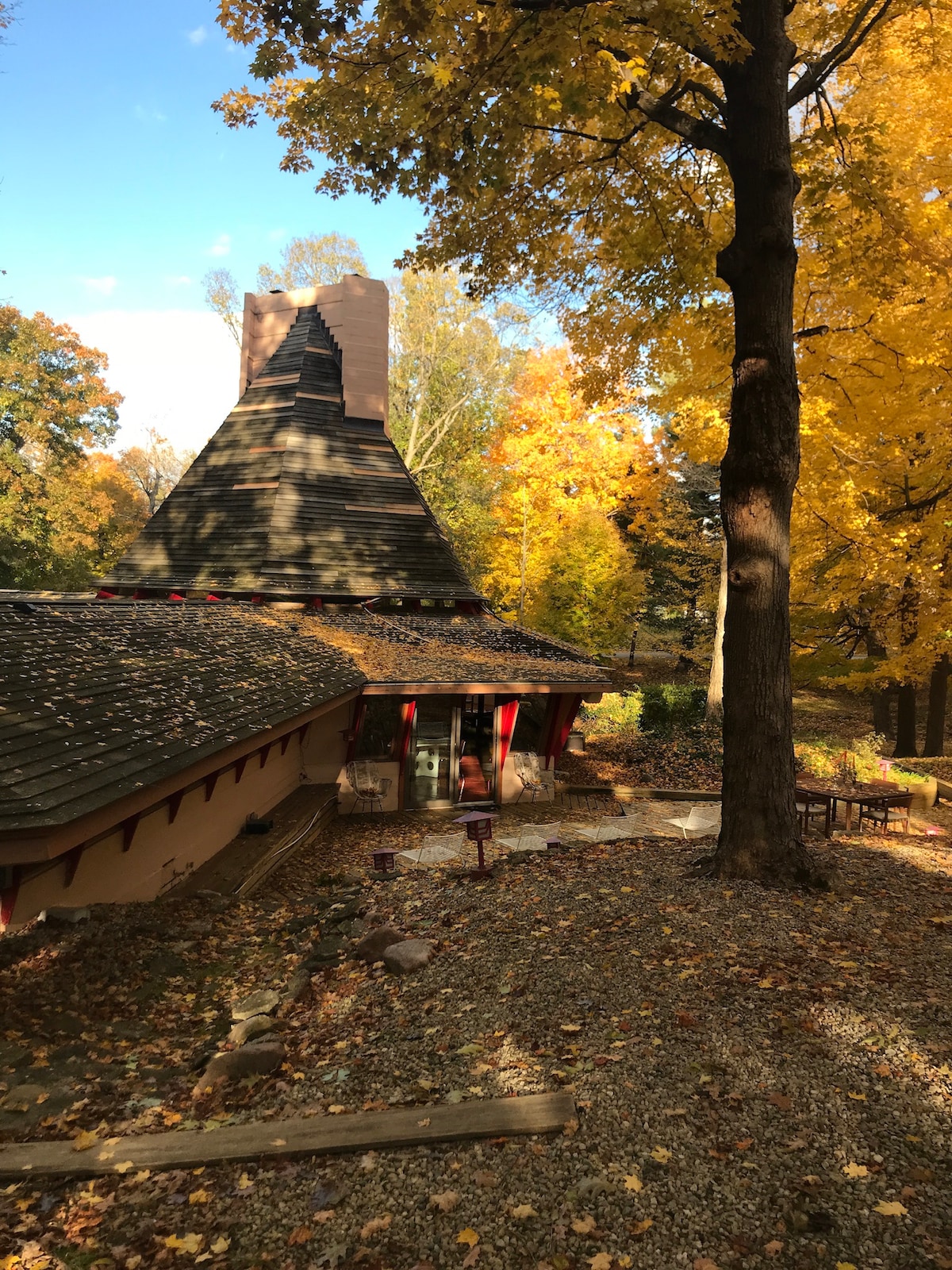 The exterior of a uniquely designed house is framed by vibrant fall foliage. The sloped roof showcases a distinctive brick chimney, while natural stone pathways meander around the structure. Outdoor seating is visible amid the colorful leaves, creating a harmonious blend with the surrounding environment.