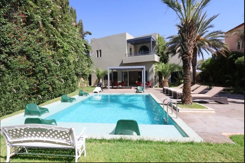 A private swimming pool is surrounded by a well-maintained garden, featuring lounge chairs on the patio. Palm trees and greenery provide a serene backdrop for the villa, which has a spacious outdoor terrace visible through large glass doors.