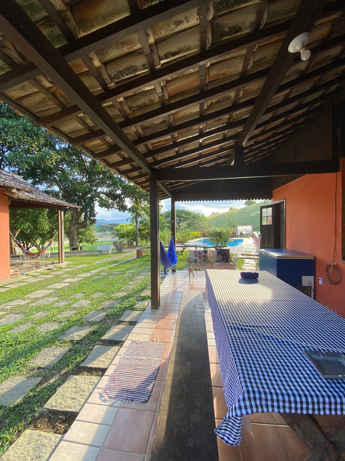A shaded outdoor area features a long table covered with a checkered tablecloth. Lush green grass is visible, with a stone pathway leading towards a view of distant hills. Blue umbrellas are placed nearby for seating under the open sky.