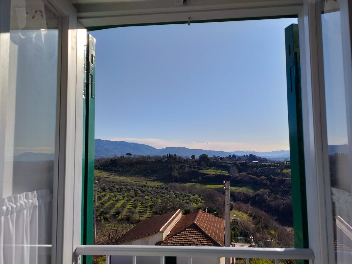 A serene view is visible through the open window, showcasing rolling green hills and the valleys below. Lush olive groves and distant mountains create a picturesque landscape under a clear blue sky.
