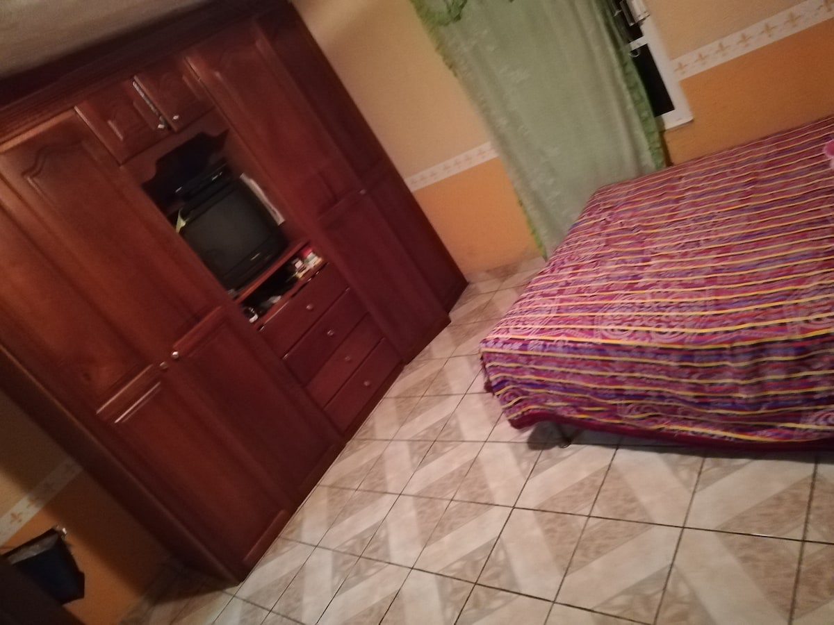 A cozy bedroom features a colorful striped bedspread on a spacious bed. A wooden wardrobe with drawers is present, alongside a small television. Light green curtains frame a window that adds natural light to the room, while tiled flooring is visible throughout.