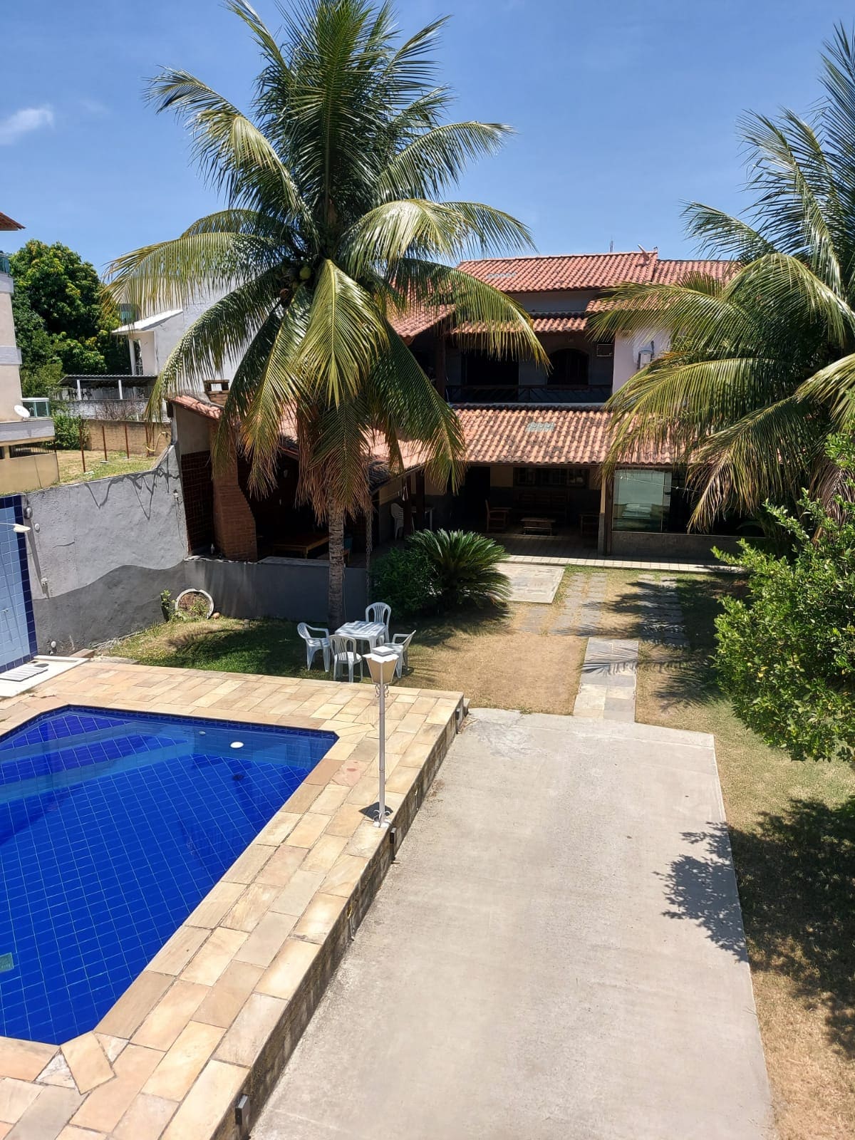 The outdoor area features a refreshing swimming pool surrounded by palm trees and a stone patio. A well-maintained lawn leads towards a spacious house with a terracotta roof. Lightweight plastic chairs are positioned nearby, complementing the tranquil atmosphere.