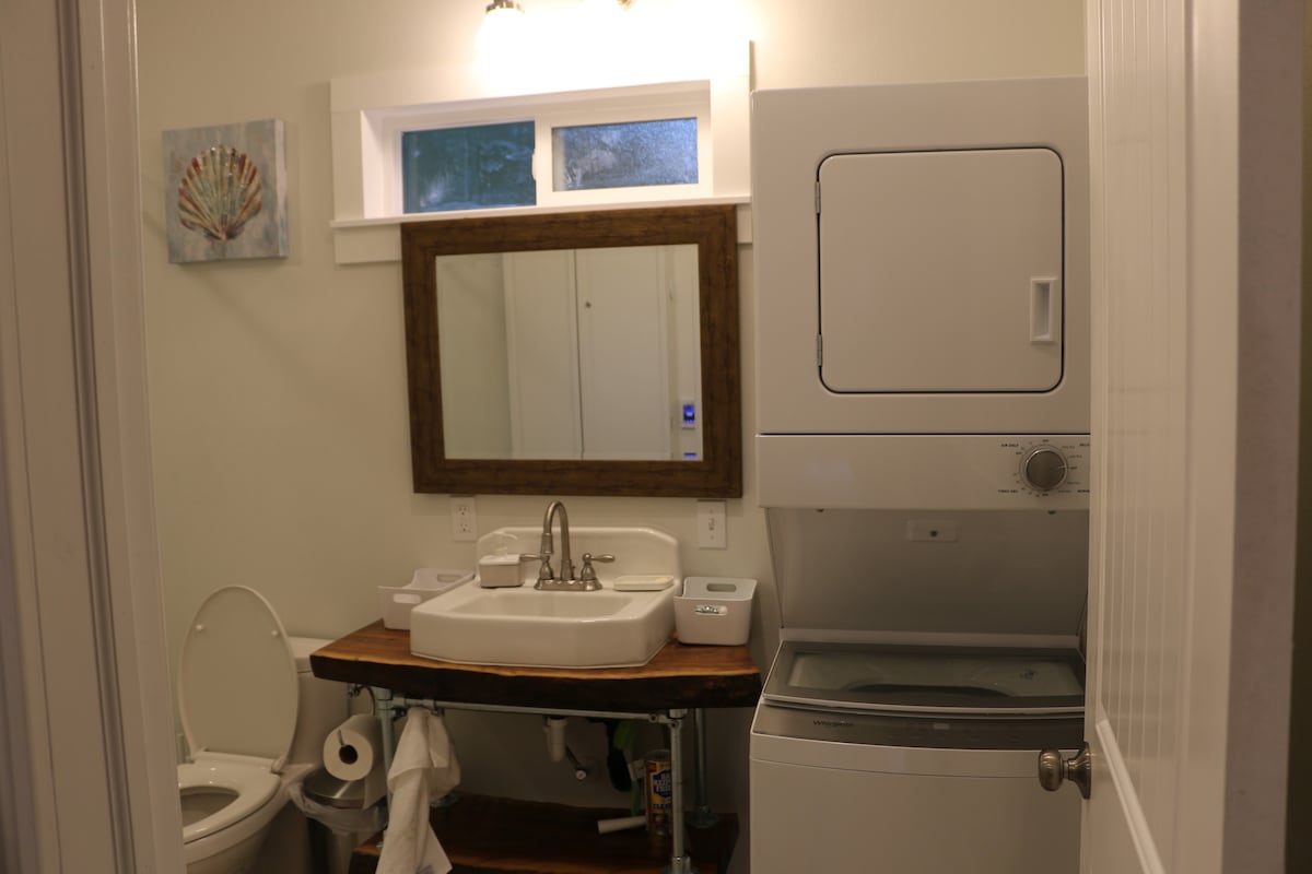 A clean and functional bathroom features a wooden countertop with a square sink and a large mirror above it. A compact washer and dryer are positioned beside the sink. Natural light filters through a window, enhancing the space's functionality.