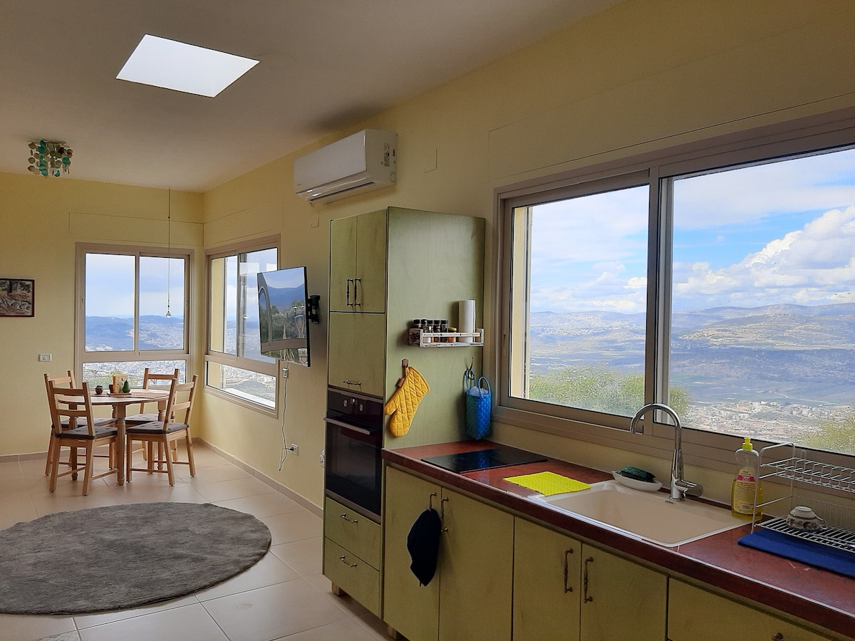 A spacious kitchen area includes a sink and stove, complemented by soft green cabinetry. Large windows offer a panoramic view of the surrounding landscape, while a dining table with chairs invites casual meals. Natural light streams in from the overhead skylight.