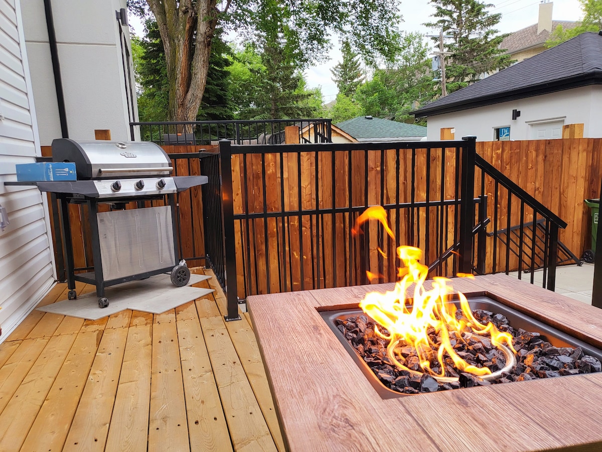 An outdoor deck area includes a stylish fire pit with vibrant flames dancing above black rocks. Adjacent to it, a grill sits on a tiled surface, ready for outdoor cooking. Surrounding wooden fencing provides privacy and seclusion.
