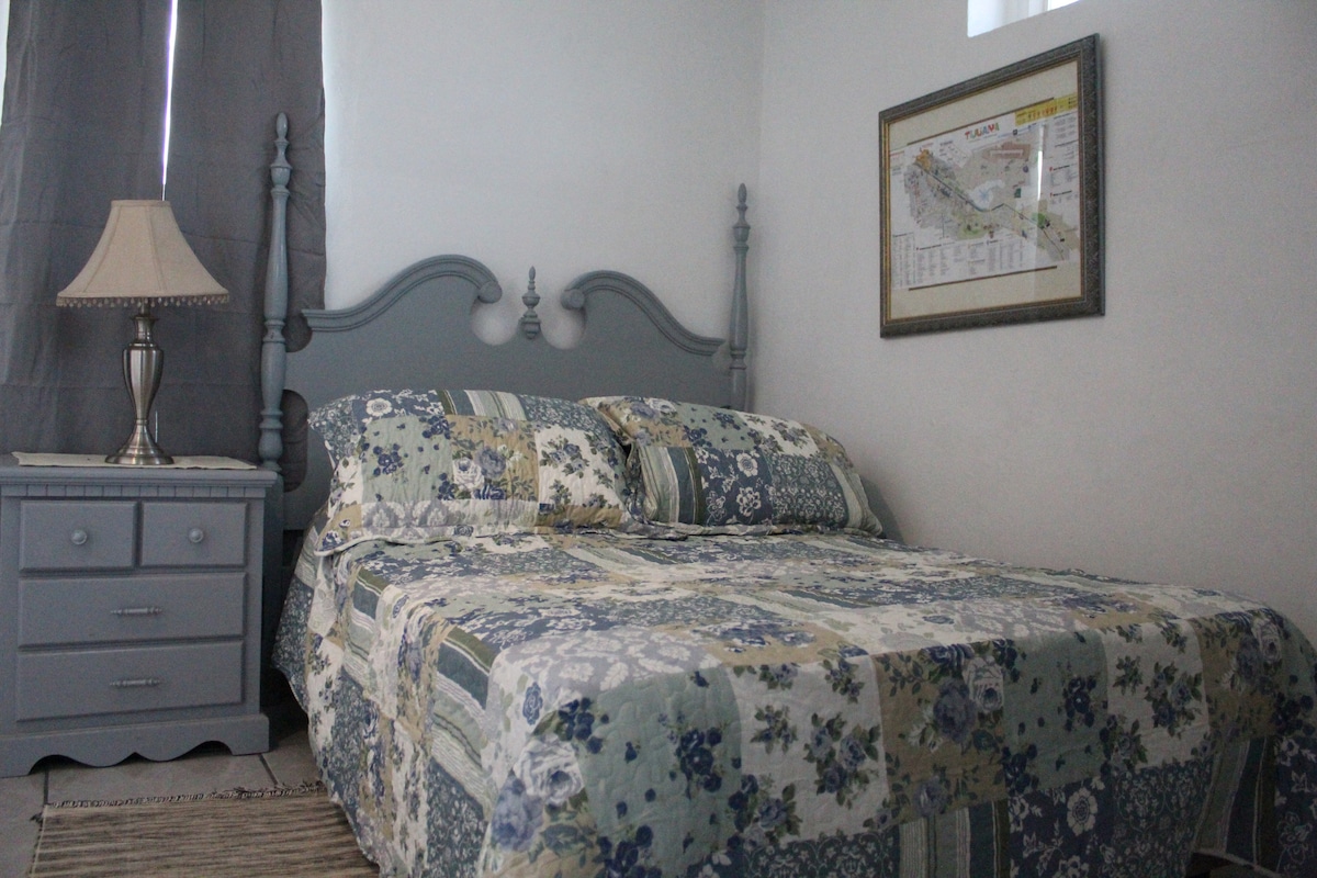 A cozy bedroom features a spacious bed with a floral patchwork quilt. A bedside table holds a lamp, providing soft illumination. Light gray curtains are drawn to the side of a window, and a map hangs framed on the wall, adding subtle decor.