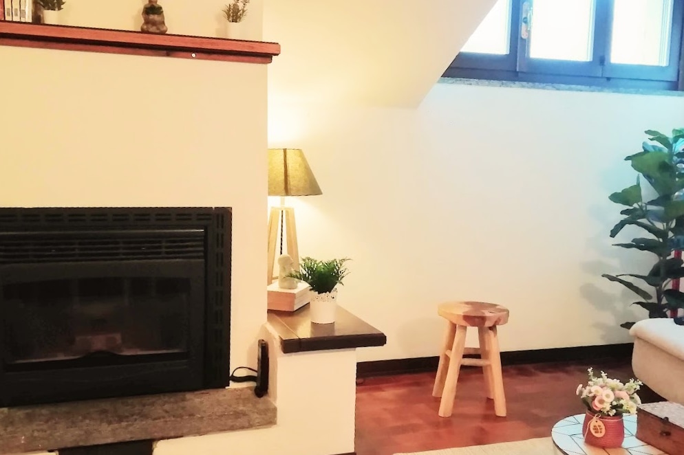 A cozy corner of the living area features a decorative fireplace surrounded by a stone ledge. A small wooden stool sits nearby, complemented by a lamp that adds subtle lighting. A potted plant is positioned against the wall, enhancing the serene atmosphere.