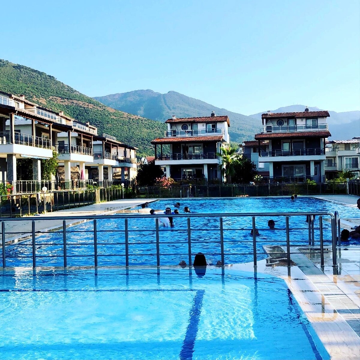 A clear, inviting swimming pool is surrounded by contemporary multi-story buildings, with lush greenery and mountains in the background. The pool area is designed for relaxation, featuring comfortable lounging spaces and gentle waves from swimmers enjoying the day.