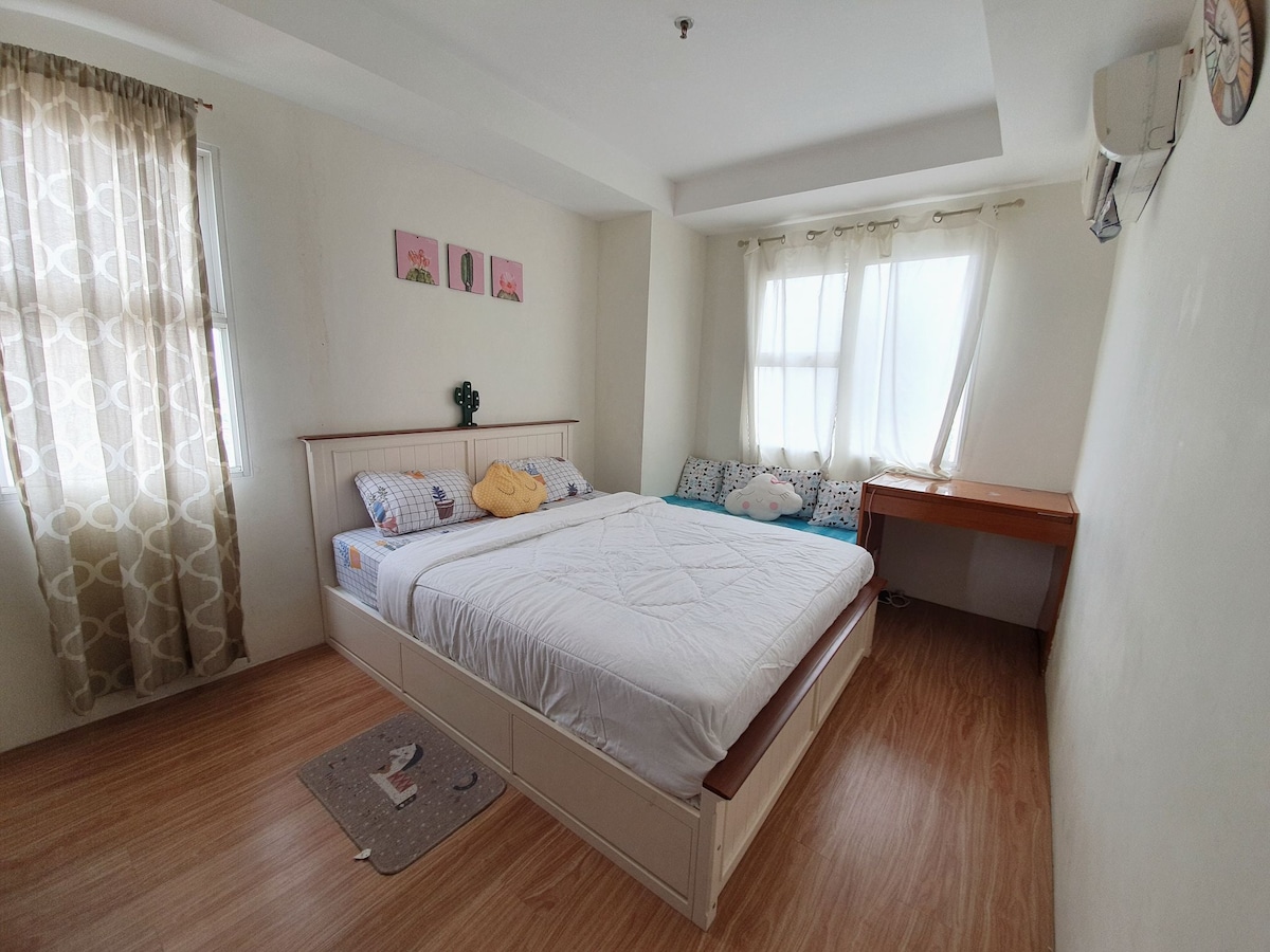 A bright bedroom features a spacious bed with a simple white comforter. Soft natural light enters through two windows, complemented by sheer curtains. A small desk is visible next to the bed, and decorative cushions add a touch of comfort to the room.