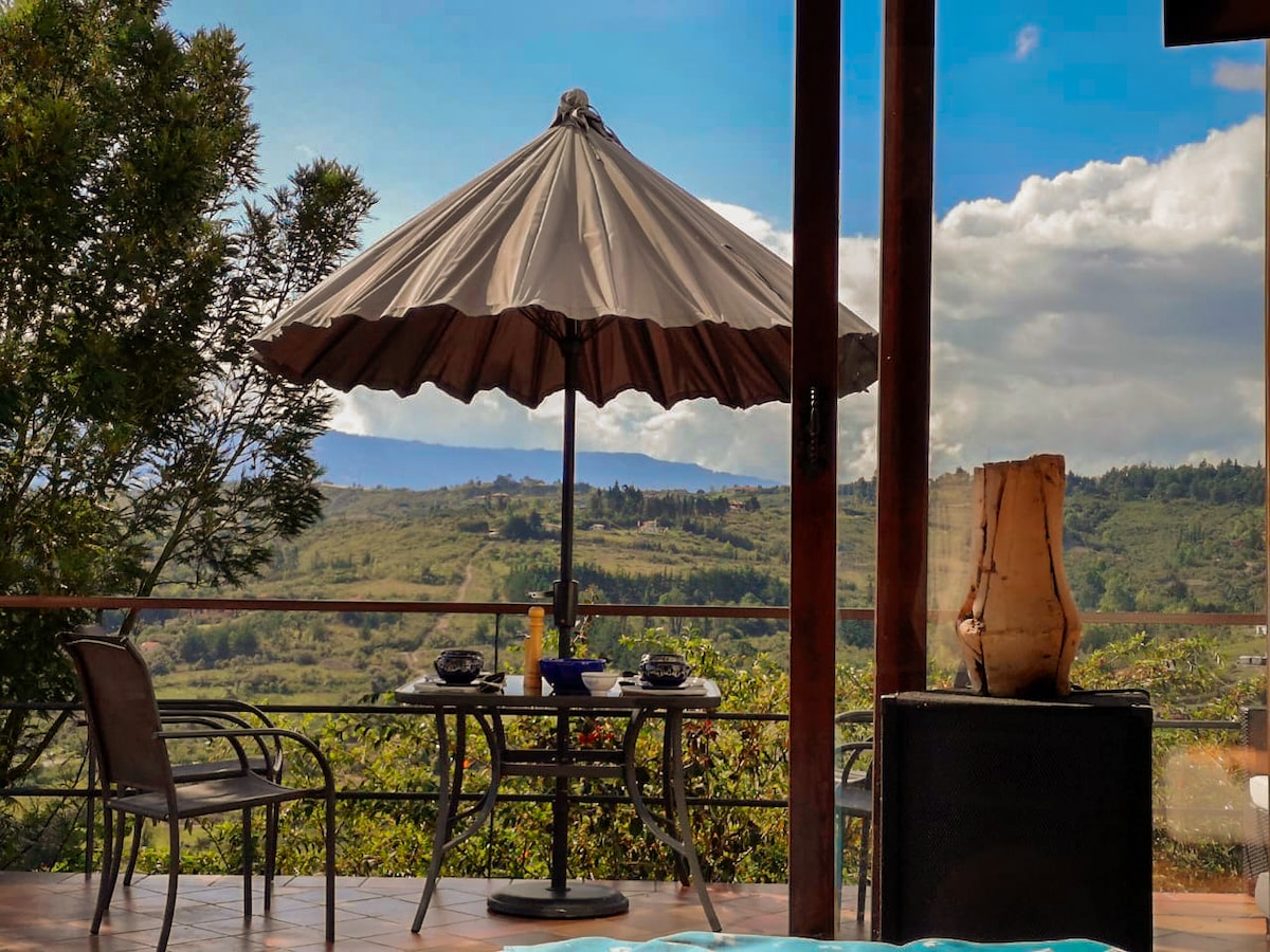 A shaded patio area features a large umbrella over a circular table, accompanied by four chairs. A serene view of rolling hills and distant mountains is visible in the background, providing a calming outdoor space for relaxation.
