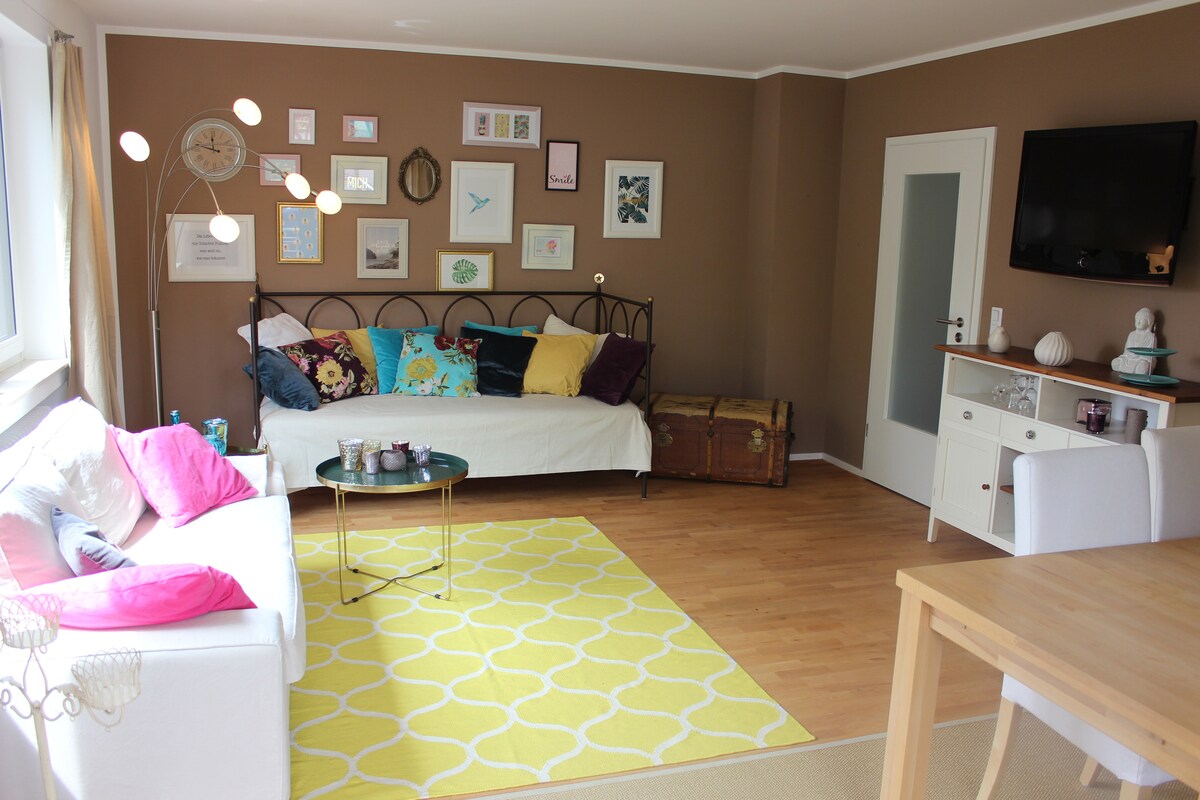 A spacious living area features a bright rug with a geometric pattern and a mix of decorative cushions on a daybed. A wooden dining table is present, along with a media console displaying a television. Natural light streams in through large windows.