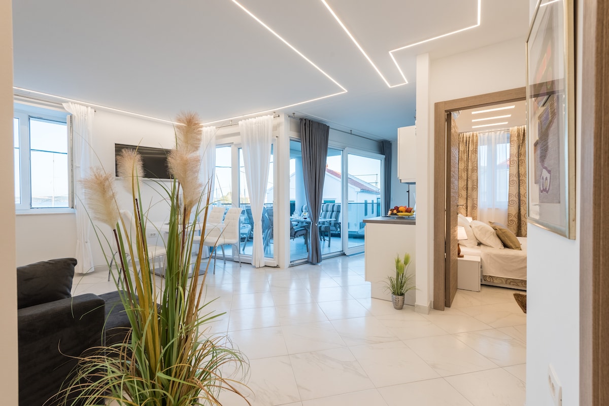 A spacious living area features modern decor and large windows, providing ample natural light. A comfortable sofa is complemented by decorative plants. An adjoining dining space is visible, with a dining table and chairs. The open layout leads to a kitchen area, enhancing convenience.