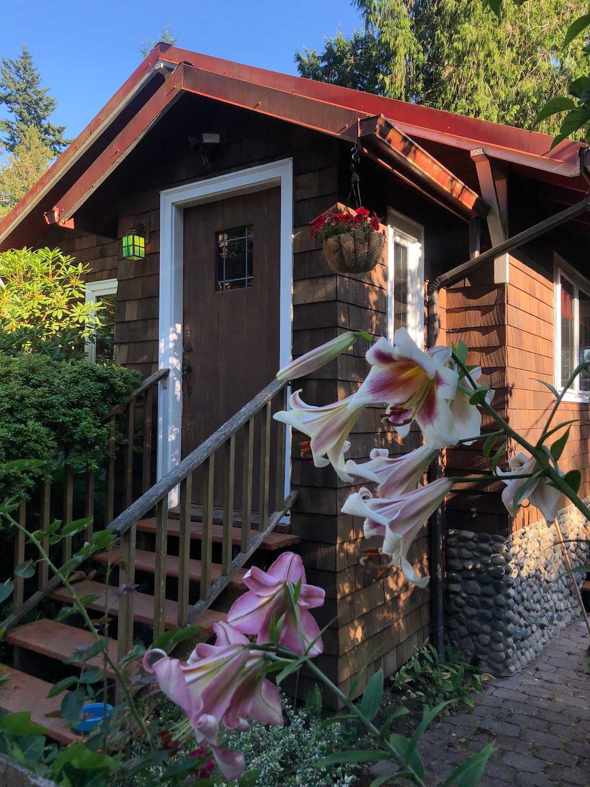 The inviting entrance to the cabin features a wooden door framed by lush greenery and blooming flowers. Steps lead up to the door, with a decorative planter hanging nearby, enhancing the natural charm of the cabin's exterior.
