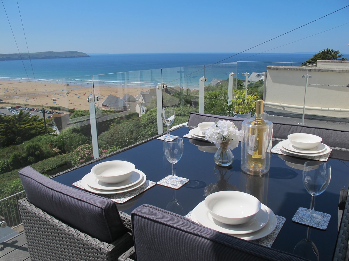 A dining table set for four is positioned on a balcony, featuring clear glass railings that offer panoramic views of the sandy beach and ocean. White plates and wine glasses are neatly arranged, with a decorative centerpiece adding a touch of elegance to the outdoor dining space.