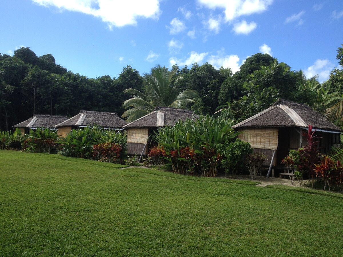 Three bungalows with thatched roofs are situated amidst lush greenery, featuring vibrant tropical plants and a well-maintained lawn. The serene natural surroundings provide a peaceful atmosphere, while the open space offers room for outdoor activities.