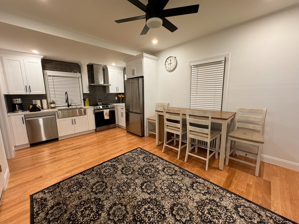 A modern kitchen features stainless steel appliances, including a gas stove and refrigerator. A dining area with a wooden table and four chairs is seen alongside a patterned area rug. Natural light enters through the window, accentuating the bright and open space.